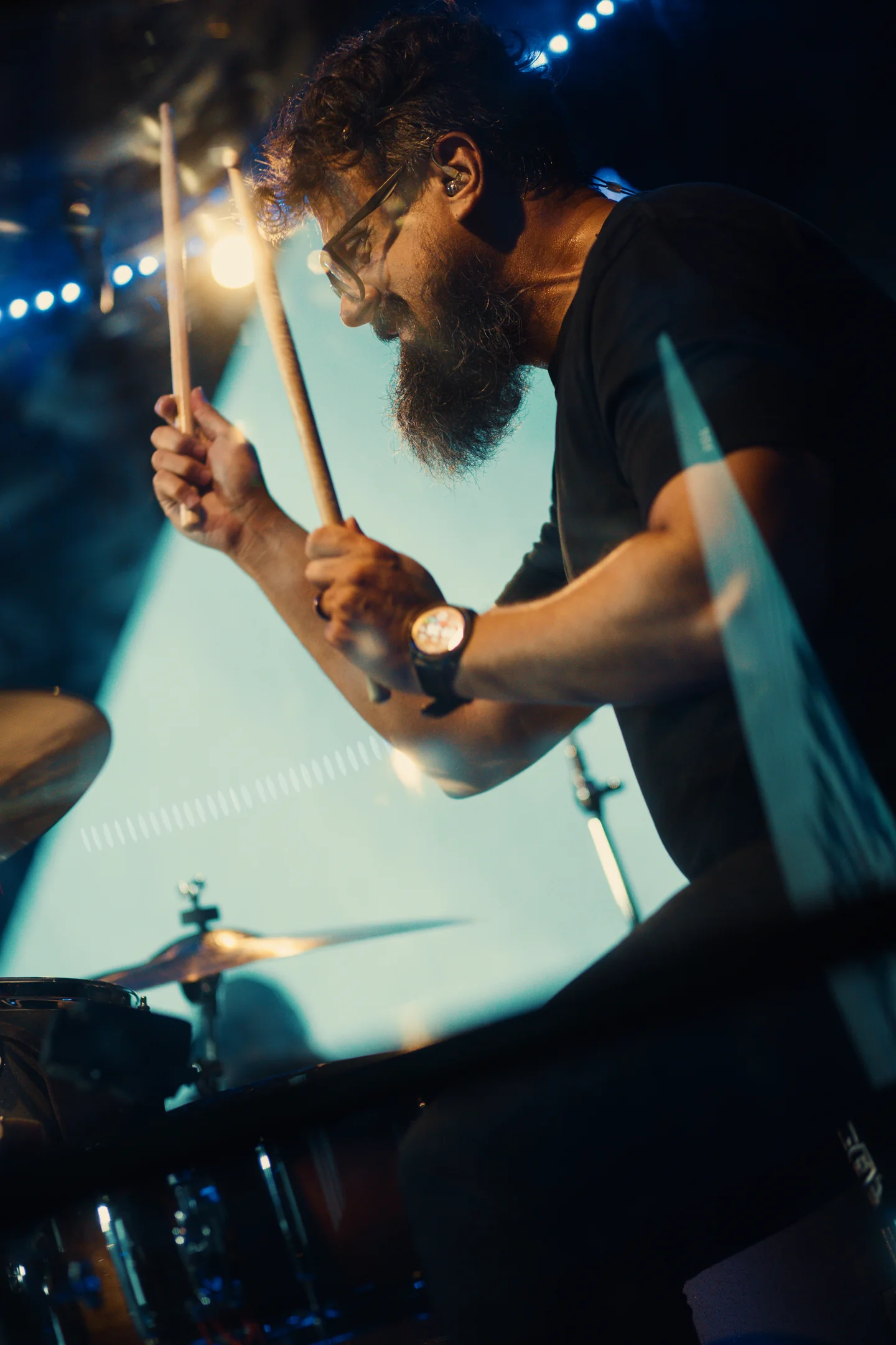 Baterista com as baquetas levantadas, entre luzes azuis e um ponto de luz quente.