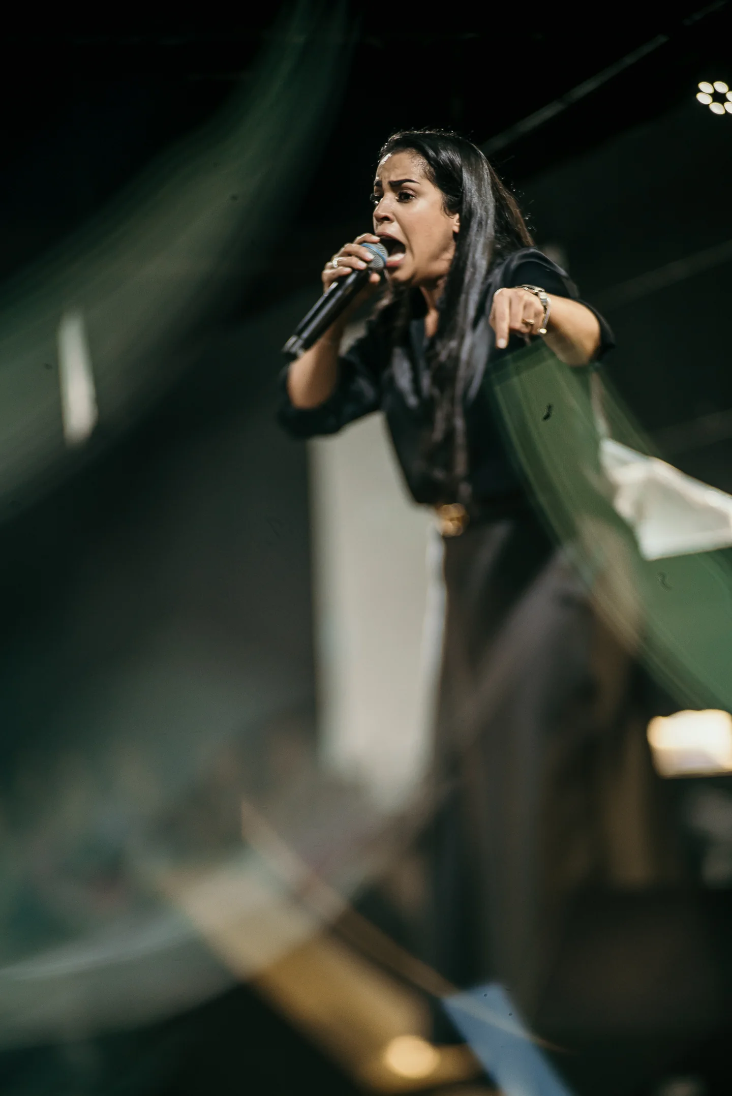 Cantora ao microfone, com a mão estendida e elementos do palco desfocados em primeiro plano.
